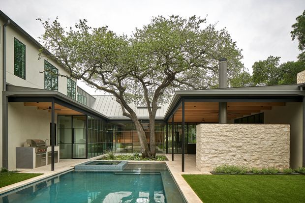 A contemporary courtyard-style residence arranged around a long swimming pool and mature central tree. The low, flat-roofed wings feature light rendered walls, extensive dark-framed glazing, metal roof sections, and warm timber-lined soffits. A textured pale stone feature wall partially screens an outdoor living area, with manicured lawn and restrained landscaping creating a refined indoor-outdoor composition.