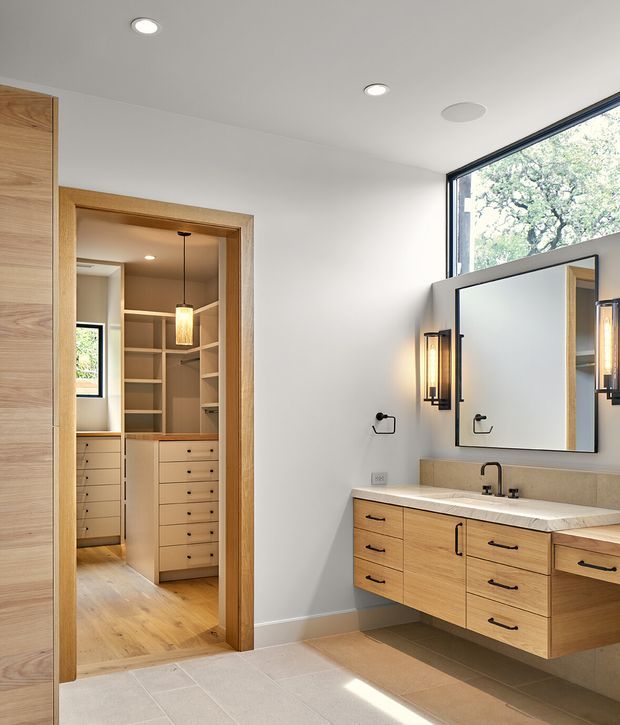 Bright contemporary bathroom with a floating light-wood double vanity, pale stone benchtop, large frameless mirror, and warm wall sconces. A walk-in wardrobe with matching timber cabinetry is visible directly through the open doorway, establishing a bedroom-suite connection. The room features pale tiled flooring, white walls, recessed ceiling lights, and a large corner window overlooking trees.