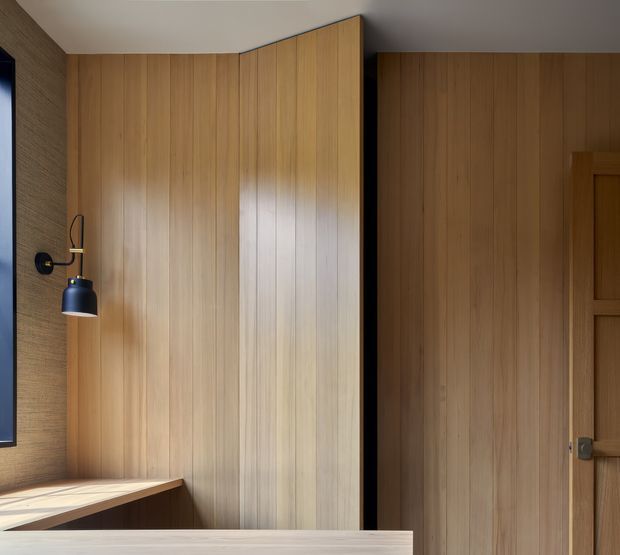 A close-up interior view dominated by warm vertical timber wall panelling and integrated timber cabinetry or a concealed door. A slim timber desk or bench runs beneath a black and brass wall sconce, with a dark-framed opening at left and a timber panel door at right. The restrained materials and clean detailing create a warm, refined contemporary interior.