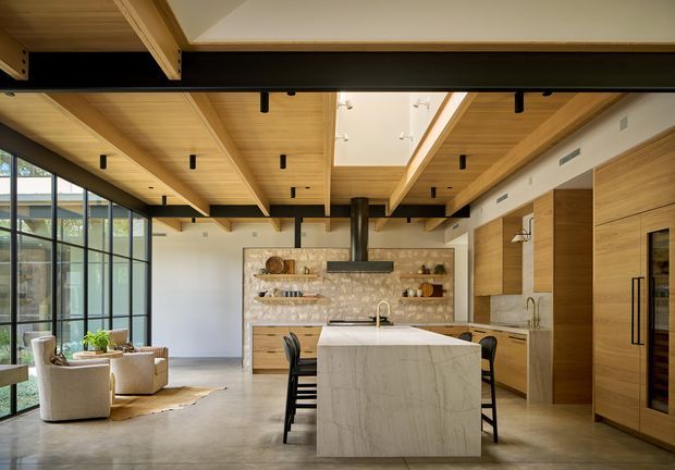 A spacious open-plan kitchen and living area with a large marble-look island, warm timber cabinetry, exposed timber ceiling beams, black steel structural framing, and full-height glazed walls. The kitchen includes integrated appliances, open shelving, pendant-free recessed lighting, and a dark rangehood. A small seating area is visible beside the glazing, and a skylight or clerestory opening brings in additional daylight.
