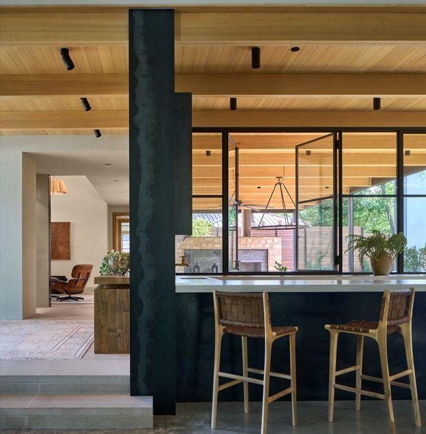 A contemporary open-plan kitchen and living area with a dark island and raised breakfast bar, two woven timber bar stools, and a large black-framed glazed opening to a landscaped outdoor courtyard. The ceiling is clad in warm timber boards with recessed and track-style spotlights. A dark sculptural structural column and matching rangehood create a strong vertical feature, while the adjoining living area contains a lounge chair and textured wall panel.