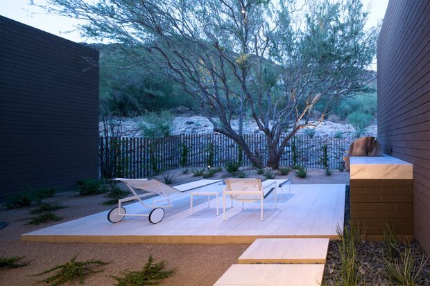 A contemporary outdoor terrace or courtyard framed by dark horizontal-clad walls, with pale concrete or stone paving, timber stepping slabs, minimalist outdoor lounge furniture, and a built-in raised bench or counter along the right wall. The space opens onto a dry, landscaped setting with native-looking trees, gravel, rocks, and a slatted privacy fence.