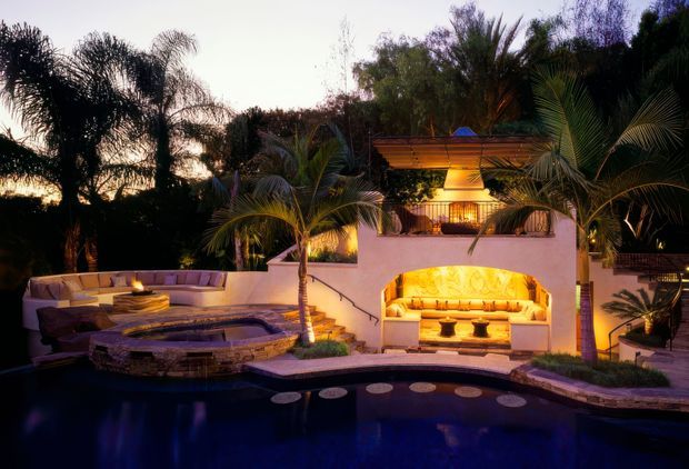 Twilight view of a luxurious tropical outdoor entertaining area centered around a swimming pool. The space features a curved built-in lounge with warm integrated lighting, a raised circular spa or firepit-like feature, a covered outdoor seating pavilion with a glowing built-in banquette, palms and dense landscaping, and arched stucco masonry detailing.