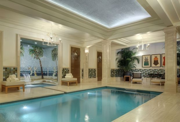 Luxurious indoor swimming pool room with a large turquoise pool, pale stone or tile surrounds, decorative classical columns, coffered ceiling, chandeliers, patterned mosaic wall tiles, tropical artwork and plants, and lounge seating. The space has a refined resort-like atmosphere with a skylight above the pool.