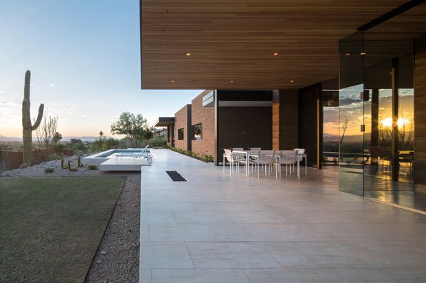 A contemporary single-storey residence viewed from a broad outdoor terrace at sunset. The home features dark brick walls, extensive floor-to-ceiling glazing, a deep cantilevered timber-lined soffit with recessed lights, and a minimal pale tiled terrace. An outdoor dining setting sits beside the glazed living area, with a linear pool and desert-style landscaped garden beyond.