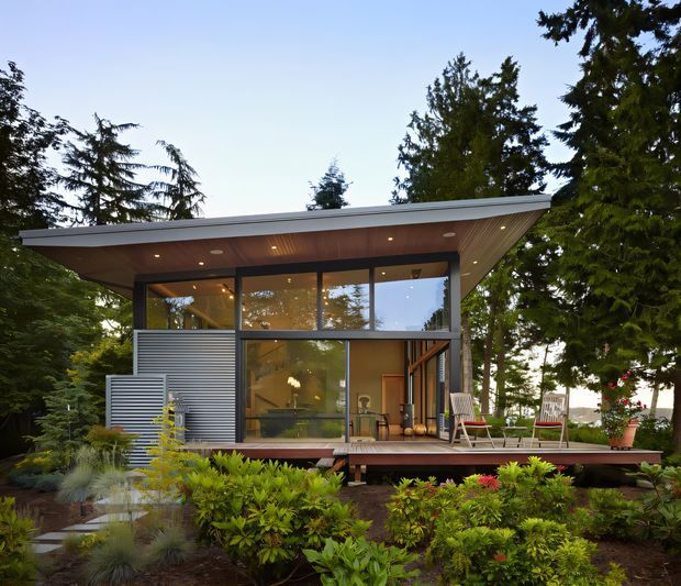 Exterior view of a compact contemporary single-storey pavilion-style house set within a landscaped garden. The dwelling has extensive floor-to-ceiling glazing, a broad low-pitched roof with exposed timber soffit, dark structural framing, corrugated metal wall cladding, and a raised timber deck with outdoor chairs. Mature trees and dense planting surround the building.