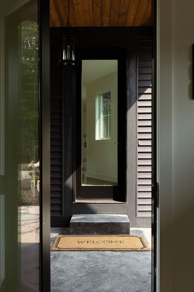 A narrow, dark and highly contrasted entryway viewed through an open glazed door. The entrance features black-painted horizontal cladding and trim, a warm timber-lined ceiling, a hanging black lantern, a raised stone or concrete threshold step, and a tan welcome mat. A full-height mirror or reflective glazed panel forms part of the inner door, reflecting a bright adjoining interior and window. Garden landscaping is visible through the side glazing.