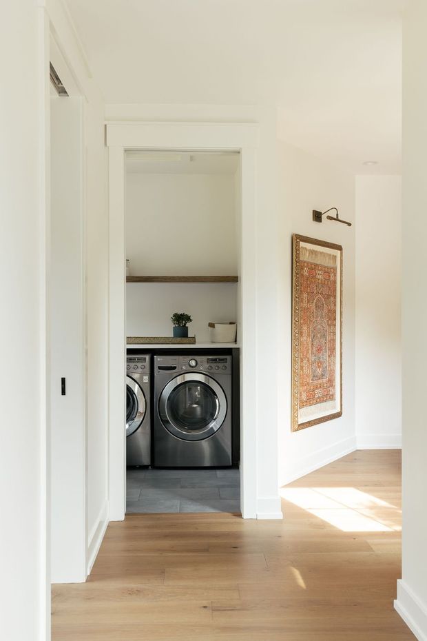 Bright, narrow interior view from a hallway into a compact laundry cupboard. The laundry contains two front-loading stainless-steel washing machines, open timber shelves, and a dark benchtop. White walls, trim, and cabinetry surround the opening, while the adjoining hallway has warm timber flooring and a framed textile artwork illuminated by a wall light.