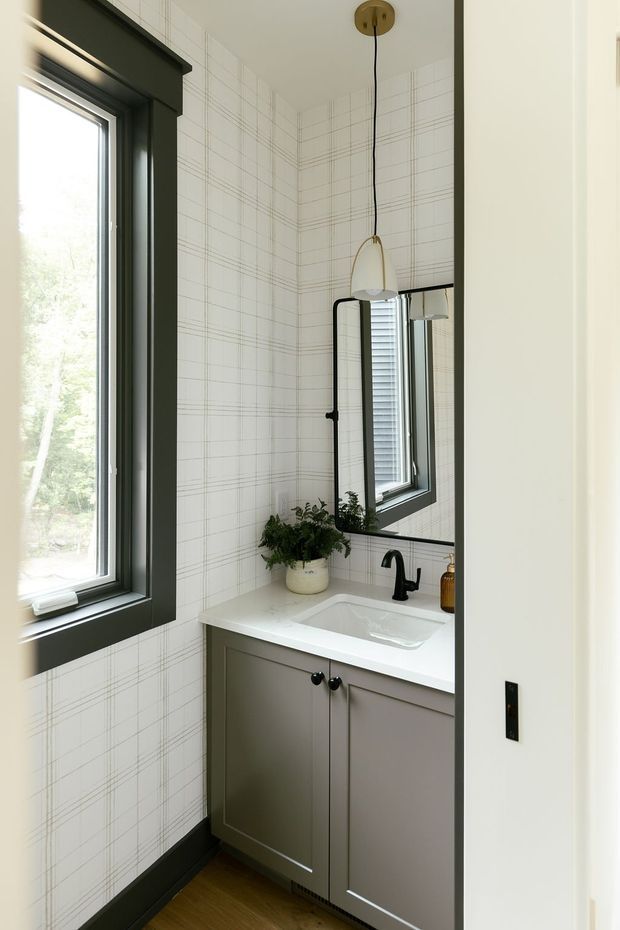 A compact, bright bathroom vanity area with a white rectangular basin set into a pale grey built-in cabinet, black tapware, and a large black-framed mirror. The walls are covered in white grid-patterned tile, complemented by dark green-black window and skirting trims, warm timber flooring, a hanging pendant light, and a small potted plant.