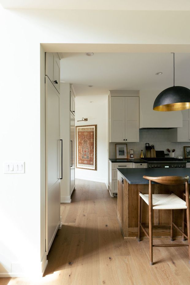 Warm, refined kitchen viewed through a wide doorway, with pale shaker-style cabinetry, a dark stone island benchtop, timber island base, light timber flooring, and a black-and-brass dome pendant. A hallway with framed textile artwork extends beyond the kitchen.