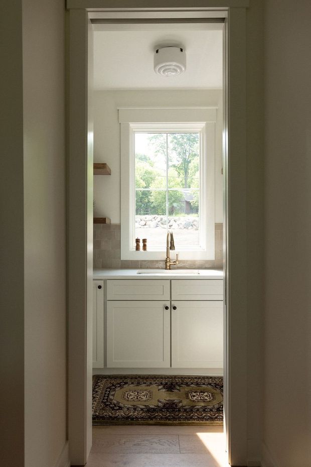 A narrow view through a doorway into a compact, traditional-style kitchen. The room has pale painted shaker cabinetry, a light stone benchtop, a tiled backsplash, a brass gooseneck tap, a white-framed multi-pane window, open timber shelves, and a patterned runner over a light timber floor. Natural light enters through the window and doorway, creating a calm, understated interior.