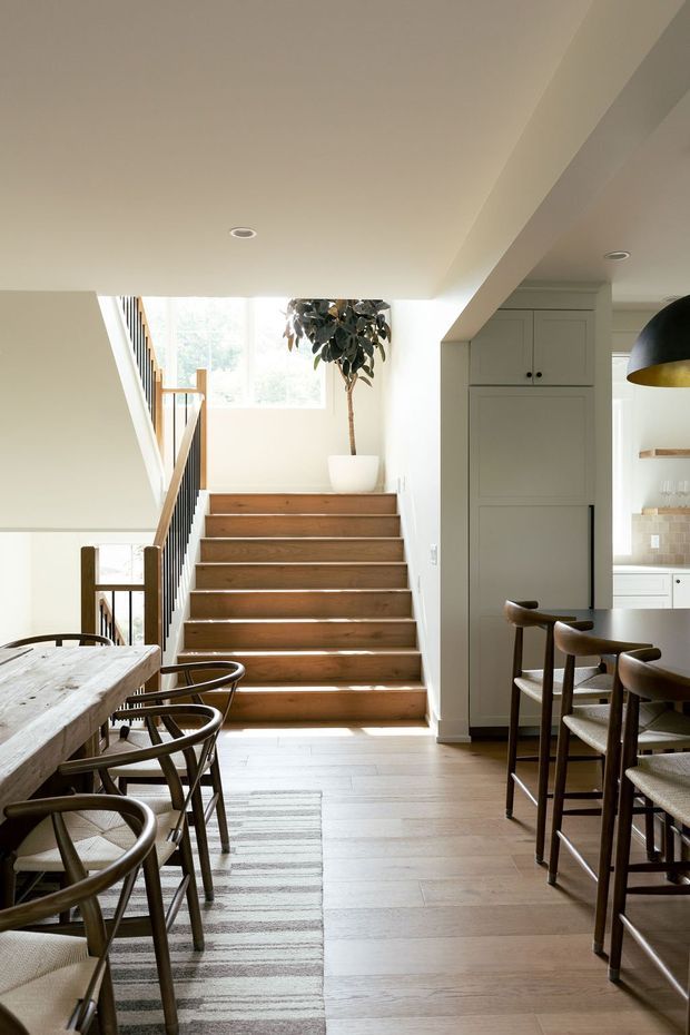 Bright open-plan interior showing a timber staircase, dining table and chairs in the foreground, and a kitchen with fixed cabinetry and tiled splashback at right. The space features warm timber flooring and stair treads, light neutral walls and cabinetry, dark-framed balustrade details, recessed ceiling lights, and a sculptural pendant over the kitchen or island area.