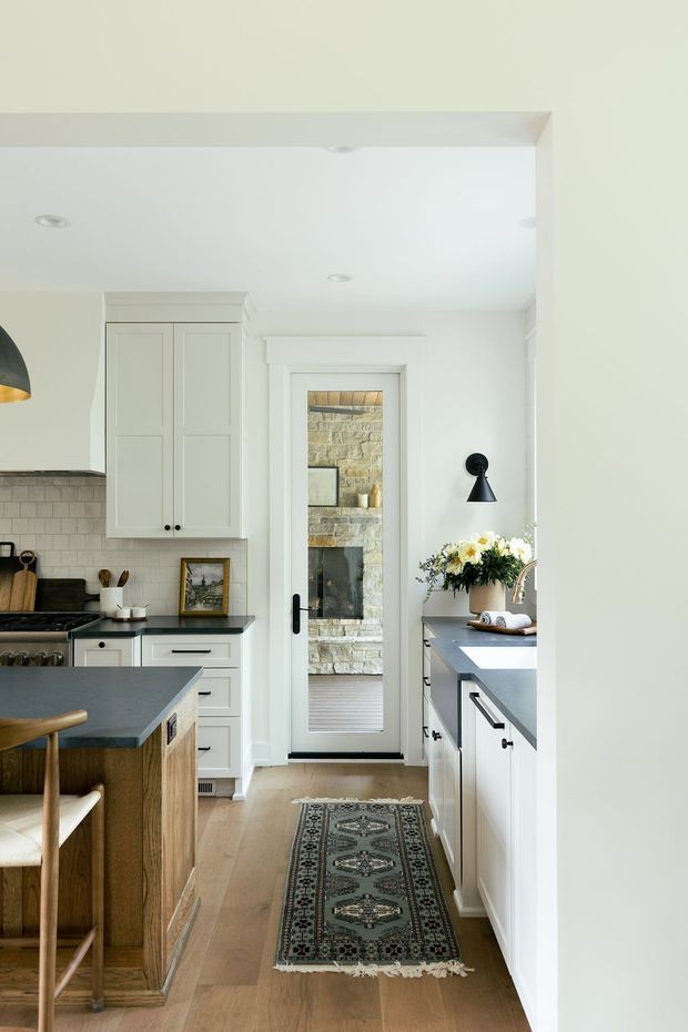Bright, narrow galley-style kitchen with a central timber island, pale paneled cabinetry, dark stone benchtops, and a light tiled backsplash. A glass door at the rear opens toward a stone-clad fireplace or adjoining living area. White walls, recessed ceiling lights, a black wall sconce, timber flooring, and a patterned runner create a refined traditional interior.