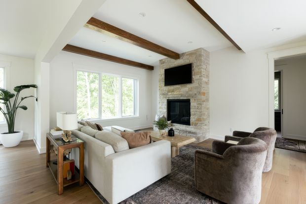 Bright, refined living room with a white sectional sofa and two plush lounge chairs arranged around a stone-clad fireplace. The room features white walls, recessed lighting, exposed dark timber ceiling beams, pale timber flooring, a large rug, and an adjacent doorway leading to another space.