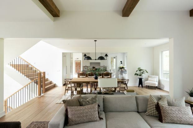 Bright open-plan living and dining area with a large grey sectional sofa in the foreground, a timber dining table and chairs beyond, and a visible kitchen at the rear. The space features light timber flooring, white walls, exposed dark timber ceiling beams, black pendant lighting, large windows, and a staircase with timber treads and slim dark balusters. The overall style is warm, contemporary, and airy.