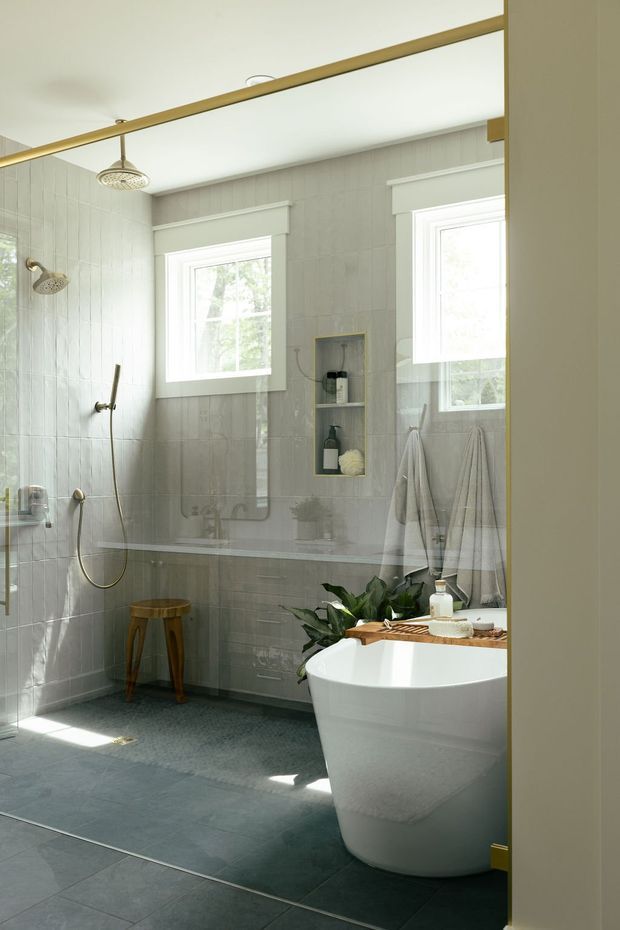 Bright, refined bathroom with a freestanding white bathtub, glass-enclosed walk-in shower, dual shower fittings, recessed wall niche, two windows, towel rails, and a small timber stool. Pale vertically textured wall tiles contrast with dark grey floor tiles. A large mirror reflects the shower and vanity area, while brass-toned mirror trim and hardware add warmth. Natural light and indoor greenery create an airy, serene atmosphere.