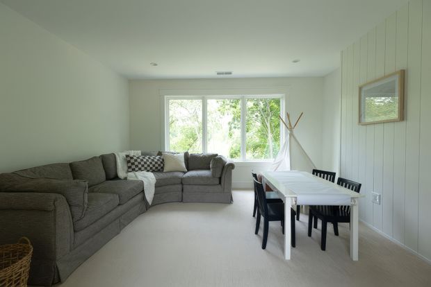 A bright, compact open-plan living and dining area. A large grey sectional sofa occupies the left side, while a white rectangular dining table with dark chairs sits beside a vertically panelled feature wall on the right. Large windows bring in abundant natural light and overlook leafy greenery. The room has pale neutral finishes, light carpet, minimal recessed lighting, and a small teepee-style decorative structure near the window.