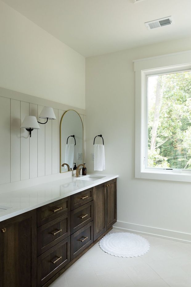 Bright, refined bathroom with a long dark timber vanity, white stone countertop, integrated double basins, brass-toned tapware and hardware, arched mirror, white vertical wall panelling, and matching wall-mounted sconces. A large white-trimmed window brings in abundant natural light, while a small round white rug sits on the pale tiled floor.