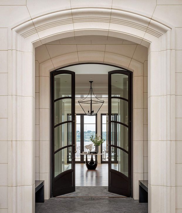 Formal architectural entrance view through a large arched opening with layered moulded detailing and pale stone wall panels. Dark framed glazed double doors stand open toward a refined interior featuring dark timber flooring, a sculptural black console or table, a geometric pendant light, and tall glazing with a distant landscape beyond. The composition is symmetrical and elegant, with strong traditional detailing and a restrained monochromatic palette.
