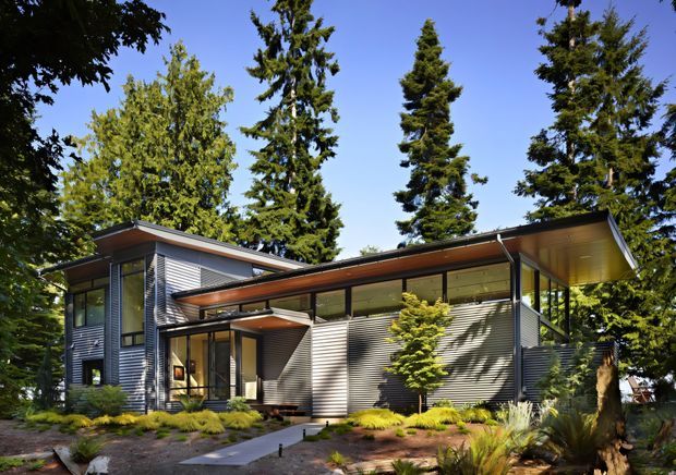 A contemporary single-storey house nestled among tall evergreen trees and landscaped garden beds. The building has a low-pitched, asymmetrical roof with broad projecting eaves, extensive dark-framed glazing, horizontal corrugated metal wall cladding, and timber-toned soffits. A paved path leads toward the recessed entry, while covered outdoor areas and layered roof forms create a strong connection to the surrounding wooded site.