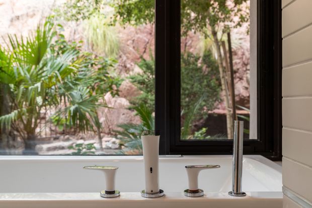 Close-up view of a freestanding or built-in white bathtub positioned beside large black-framed windows, with a chrome faucet and separate chrome hot and cold controls on the tub rim. The windows overlook lush tropical landscaping and natural rock, while horizontal light-coloured wall cladding is visible at right. The scene has a clean, modern bathroom aesthetic with strong indoor-outdoor connection.