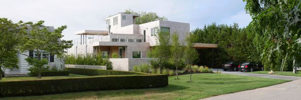 Wide exterior view of a contemporary multi-level residence set within a landscaped suburban garden. The house features pale horizontal cladding, light brick or masonry walls, projecting rectangular volumes, large glazed openings, flat roof forms, and a covered carport or garage area at right. Manicured hedges, lawn, and mature trees frame the property.