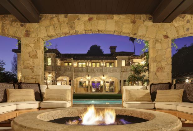 A luxurious covered outdoor lounge opening onto a courtyard pool and a large illuminated stone residence. The space features built-in cream upholstered seating arranged around a large circular stone fire pit, substantial rough-cut stone columns and architrave, dark timber ceiling beams and panelling, and warm evening lighting. The Mediterranean-style residence beyond has stone cladding, arched openings, balconies and a tiled roof.
