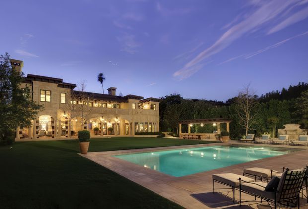 Twilight view of a large traditional-style residence with warm illuminated masonry façades, arched openings, landscaped grounds, and a rectangular swimming pool with surrounding paved terrace and outdoor lounge furniture. Dense trees frame the property, while a pergola or covered outdoor structure sits beyond the pool.