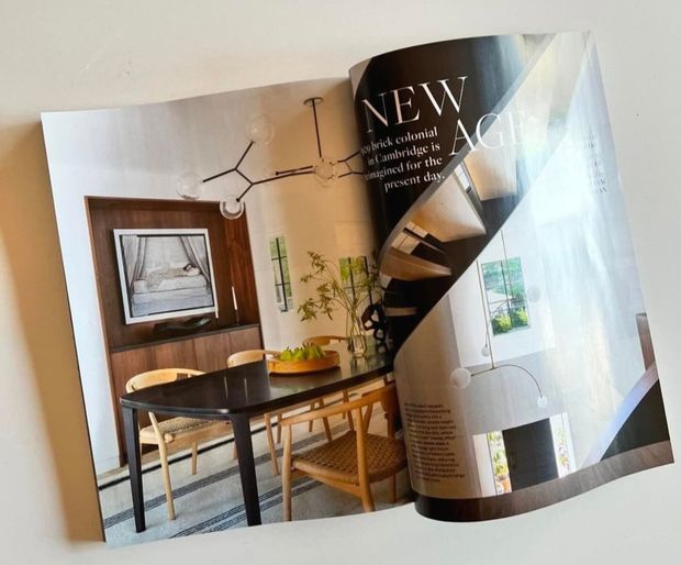A close-up photograph of an open architecture and interiors magazine spread. The visible project features a refined dining area with a dark rectangular dining table, woven timber dining chairs, warm timber wall joinery, a framed artwork or television, and sculptural pendant lighting. The opposite page shows a dramatic curved staircase or spiral stair with dark treads, a tall bright window, and pale interior surfaces. The image is primarily editorial print content rather than a direct room photograph.