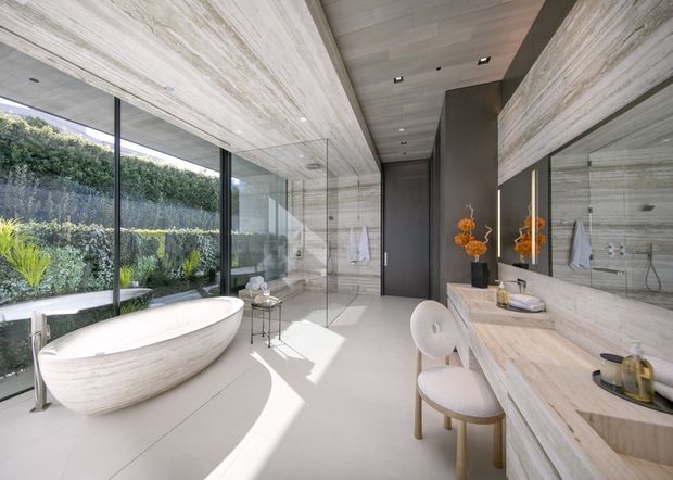 Luxurious contemporary bathroom with a freestanding oval bathtub beside full-height glazing, a long double vanity with integrated basins, large mirrors, and a walk-in shower. Light stone-look wall surfaces and ceiling create a refined, spa-like interior, with a dark timber-toned door and subtle orange floral accents. The room opens visually onto a landscaped garden through the glazing.