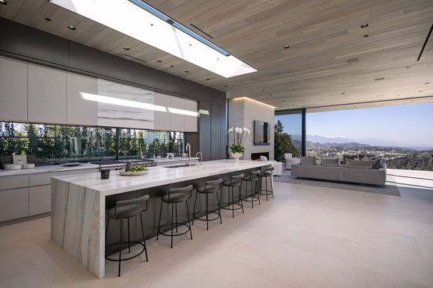 A spacious, high-end open-plan kitchen and living area with a long stone island accommodating multiple bar stools, sleek grey built-in cabinetry, integrated appliances, and a large skylight. Pale timber ceiling boards, dark-framed floor-to-ceiling glazing, and expansive views over a hilly urban landscape create a bright, contemporary interior. A minimalist living zone with a sectional sofa is visible beyond the kitchen.