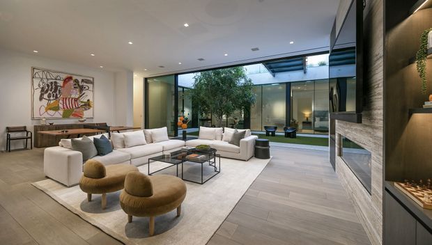 A spacious contemporary living room arranged around a large white sectional sofa and glass coffee tables, with two rounded tan lounge chairs in the foreground. The room features pale wood-look flooring, a large area rug, recessed ceiling lighting, expansive floor-to-ceiling glazing, and a central landscaped courtyard visible beyond the glass. A built-in dark timber feature wall with shelving and a stone-faced fireplace occupies the right side, while artwork and a console sit along the left wall.