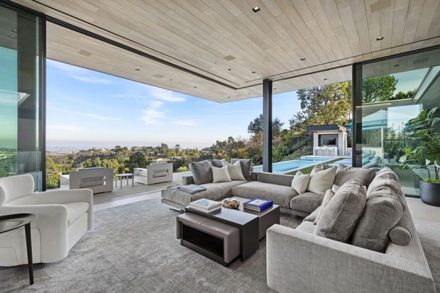 A spacious, high-end open-plan living room with a large sectional sofa, two white lounge chairs, a dark rectangular coffee table, and a pale textured rug. Full-height glazed sliding doors open the room to a covered outdoor terrace with a swimming pool and expansive landscaped hillside views. The room features a light timber slatted ceiling with recessed lighting, dark slender structural columns, and extensive floor-to-ceiling glazing.