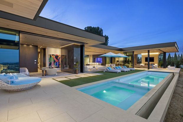 A luxurious modern single-storey residence arranged around a rectangular swimming pool and expansive paved terrace. The image shows broad flat roof planes, floor-to-ceiling glazing, dark exterior walls, warm timber soffits, recessed lighting, and an open-plan indoor-outdoor living area with lounge furniture, umbrellas, and poolside seating at dusk.