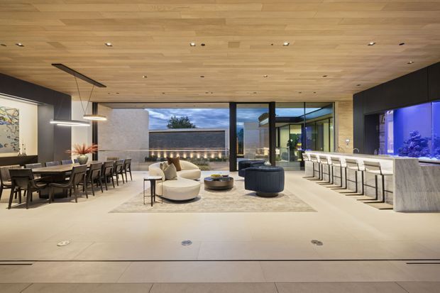 Expansive open-plan contemporary interior combining a dining area, lounge, and bar. The space features a warm timber-lined ceiling, dark charcoal walls, pale stone or concrete flooring, extensive glazing, sculptural lounge seating, a long dining table, and a stone bar with stools. A large illuminated aquarium forms a striking feature on the right.