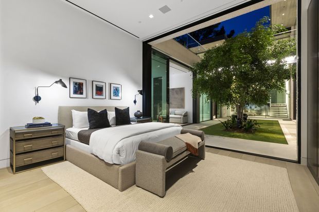 A refined contemporary bedroom with a large upholstered bed, neutral bedding, bedside cabinetry, wall-mounted reading lights, and framed artwork. Full-height black-framed glazing opens the room visually to an enclosed internal courtyard with a mature tree, lawn, and architectural circulation beyond. The room has pale timber flooring, a light textured rug, white walls, and a calm neutral palette.