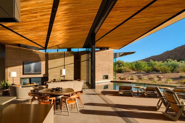 A sophisticated open-plan living and dining area extending onto a broad outdoor terrace. The space features a warm timber-lined roof with exposed dark structural beams, full-height retractable glazing, timber-toned wall panels, a fireplace and television wall, a round dining table, and outdoor loungers overlooking a dry, mountainous landscape. Strong indoor-outdoor connection, expansive glazing, and deep roof overhangs define the composition.