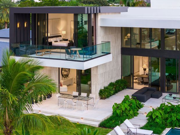 A contemporary multi-level residence viewed from the garden, featuring strong horizontal rooflines, extensive dark-framed glazing, pale stone or concrete wall surfaces, and multiple outdoor living terraces. A glazed upper balcony contains lounge seating and a small dining setting, while the lower terrace includes outdoor furniture, planting, and integrated step lighting. Tropical palms and dense greenery surround the modern house, with interior living and dining areas visible through the glazing.