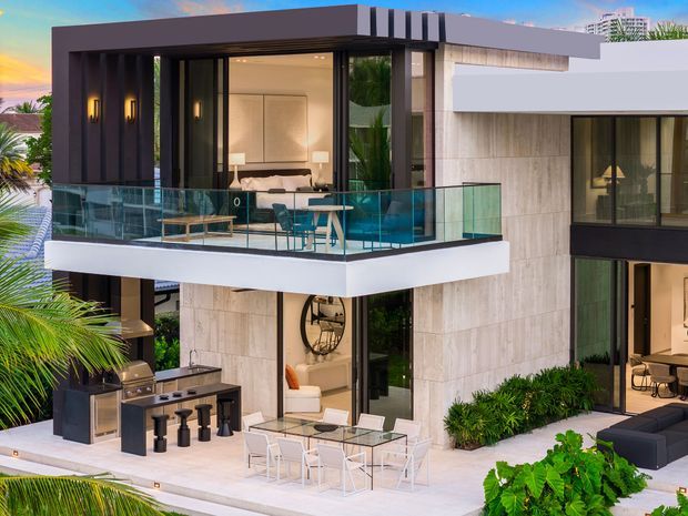 Contemporary multi-level luxury residence viewed from the exterior, featuring strong geometric volumes, dark metal framing, pale stone-clad walls, extensive glazing, and a projecting balcony with clear glass balustrades. The balcony contains an outdoor dining setting, while the lower terrace includes a built-in outdoor kitchen and dining area. Tropical planting surrounds the house, and a bedroom is visible through the upper-level glazing.