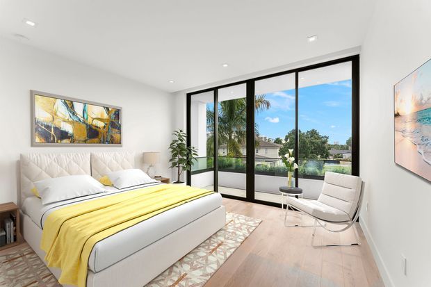 Bright contemporary bedroom with a low-profile upholstered bed, white bedding and a vivid yellow throw. The room has pale timber flooring, white walls and ceiling, recessed downlights, black-framed floor-to-ceiling glazing, a light lounge chair, artwork, a small side table, and indoor plants. The glazing overlooks greenery, palm trees, and neighbouring homes.