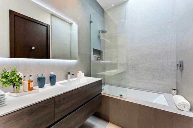 Contemporary bathroom with a double vanity in dark wood joinery, white integrated basins and benchtop, a large illuminated mirror, and a glass-enclosed shower over a built-in bathtub. The room has light grey textured wall finishes, recessed lighting, and a clean, refined fit-out.