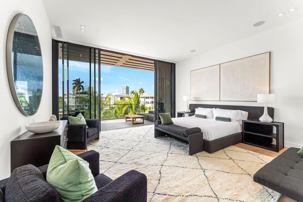 Bright, spacious contemporary bedroom with a large upholstered bed, dark charcoal lounge chairs, floating bedside tables, oversized abstract wall panels, and a large patterned rug. Full-height black-framed glazing opens to a sunny tropical outdoor view, with a timber slatted overhead detail at the opening. The room has a refined, resort-like feel with white walls and ceiling, pale bedding, and muted green accents.