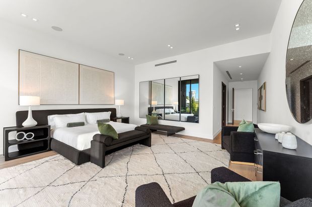Bright contemporary bedroom with a large upholstered bed, black bedside tables, a dark bench at the foot of the bed, and a large pale rug with black linear patterning. Full-height mirrored panels reflect a seating area and outdoor greenery, while a hallway and built-in dark console are visible to the right. The room has a clean white palette with muted green and black accents, recessed ceiling lighting, and minimalist furnishings.