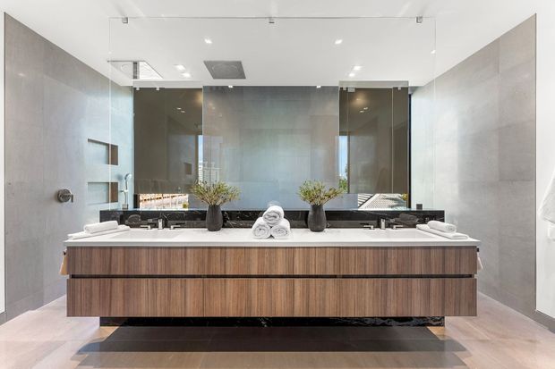 A spacious contemporary bathroom featuring a long floating double vanity with dark timber-grain cabinetry, a white stone benchtop, twin integrated basins and wall-mounted tapware. A large frameless mirror spans the rear wall, reflecting the room and windows, while grey stone-look wall and floor finishes, recessed shelving, concealed shower fittings and minimalist ceiling lighting create a refined, hotel-like setting.
