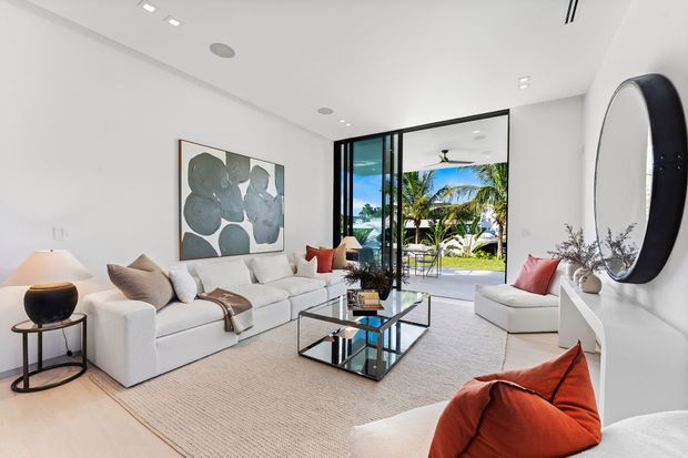 Bright contemporary living room with white walls and sofas, a large abstract artwork, glass coffee table, sculptural oval wall mirror, and warm terracotta accent cushions. Floor-to-ceiling black-framed glazing opens to a lush tropical outdoor area with a pool-like blue view beyond. The room has a high, clean-lined ceiling with recessed lighting and a calm, refined atmosphere.