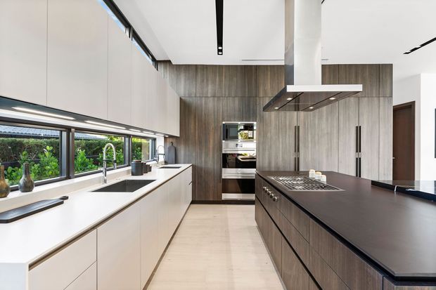 Contemporary galley-style kitchen with a large central island, dark wood-grain cabinetry, white integrated wall cabinetry, a dark benchtop, stainless-steel appliances, and a stainless-steel rangehood. A long horizontal window brings in garden views and natural light. The space has clean lines, concealed storage, and linear recessed ceiling lighting.