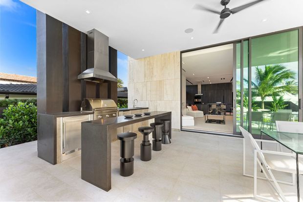 A bright, open-air contemporary outdoor kitchen and entertaining area. The space features a substantial dark stone or metal island with integrated bar seating, built-in stainless-steel barbecue cabinetry, a tall extraction hood, pale large-format tiled flooring, and a glass dining table with white chairs. Full-height sliding glazing opens to an adjacent living room, while tropical planting is visible beyond. A ceiling fan and recessed lighting complete the covered patio setting.