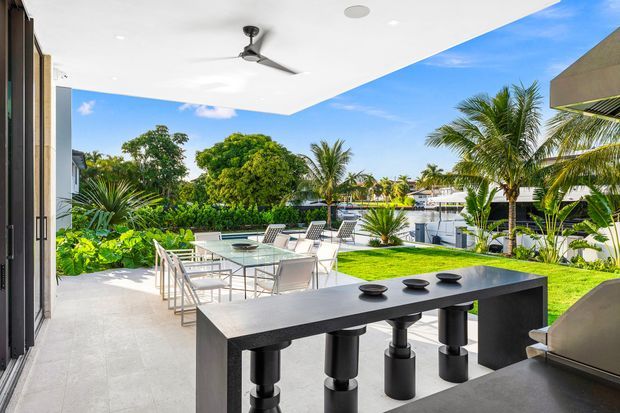 A bright contemporary outdoor entertaining area beneath a broad covered roof, with a dark built-in outdoor kitchen or bar counter in the foreground and a glass dining table with chairs beyond. The space opens onto a manicured lawn and lush tropical landscaping, with palm trees and neighbouring buildings visible under a clear blue sky.