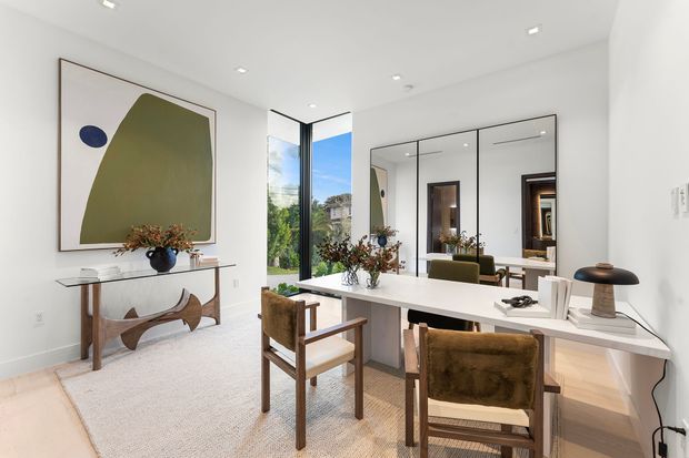 Bright contemporary dressing and study area with a long white built-in desk or vanity, two wooden-framed upholstered chairs, full-height mirrored cabinetry, pale timber flooring, and a slim console table beneath an abstract artwork. Floor-to-ceiling corner glazing brings in abundant daylight and views of greenery. The room has clean white walls, recessed ceiling lights, restrained furnishings, and warm natural wood accents.
