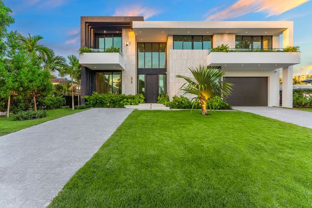 Exterior front elevation of a large contemporary two-storey residence, featuring a strong rectangular form, flat rooflines, floor-to-ceiling glazing, projecting balconies, pale stone or concrete-look facade panels, dark vertical cladding, and an integrated garage. The home is set within a manicured tropical landscape with broad lawns and a concrete driveway.